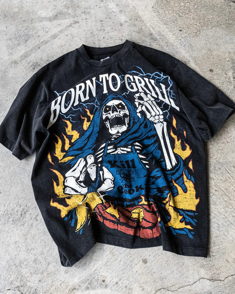 Flatlay of a vintage wash black shirt with a screen printed graphic of a skeleton grilling a steak on a BBQ. The skeleton chef wears a blue apron that says ‘Kill the Cook’ and is surrounded by yellow flames and lue lightning. Text reads ‘Born To Grill’. Skeleton chef, steak, chef tee, chef shirt