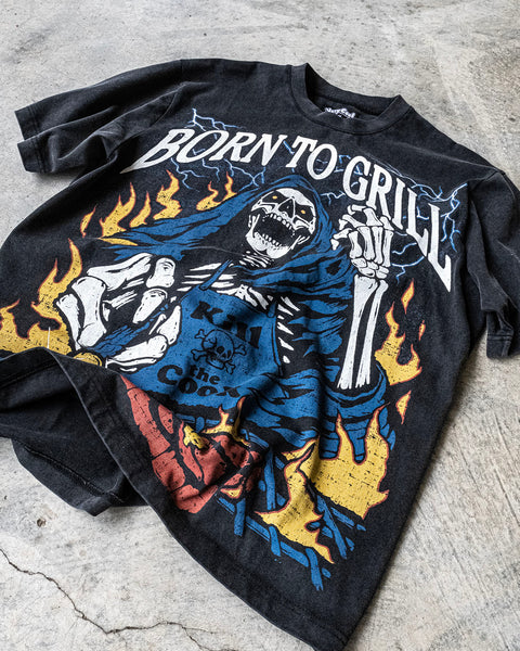 Flatlay of a vintage wash black shirt with a screen printed graphic of a skeleton grilling a steak on a BBQ. The skeleton chef wears a blue apron that says ‘Kill the Cook’ and is surrounded by yellow flames and lue lightning. Text reads ‘Born To Grill’. Skeleton chef, steak, chef tee, chef shirt