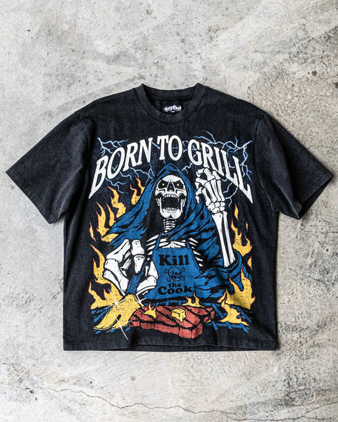 Flatlay of a vintage wash black shirt with a screen printed graphic of a skeleton grilling a steak on a BBQ. The skeleton chef wears a blue apron that says ‘Kill the Cook’ and is surrounded by yellow flames and lue lightning. Text reads ‘Born To Grill’. Skeleton chef, steak, chef tee, chef shirt