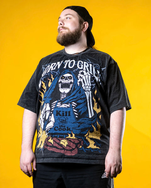 Chad from Cold Ones stands against a yellow backdrop wearing a vintage wash black shirt with a screen printed graphic of a skeleton grilling a steak on a BBQ. The skeleton chef wears a blue apron that says ‘Kill the Cook’ and is surrounded by yellow flames and lue lightning. Text reads ‘Born To Grill’. Skeleton chef, steak, chef tee, chef shirt